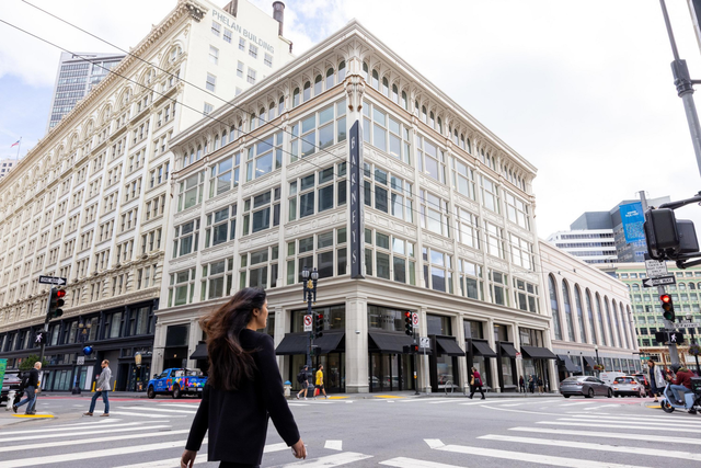 A historic corner building with large windows and black awnings stands at a busy city crosswalk with several pedestrians and cars around.